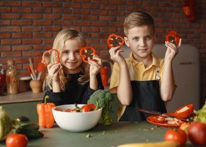 Two children holding capsicum slices while preparing vegetables in a cozy kitchen setting.