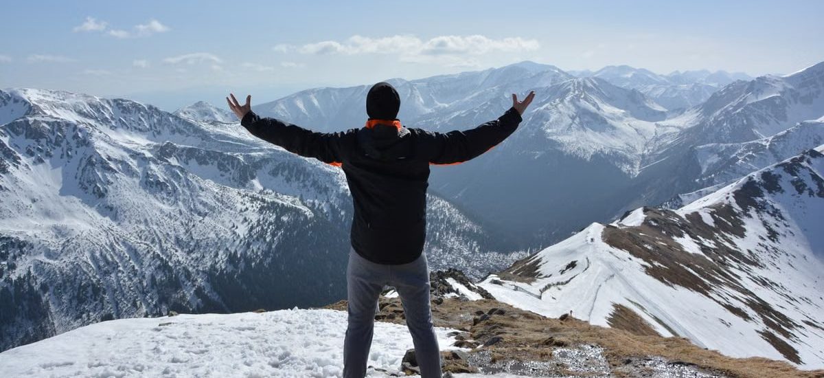A man stands on a snowy mountain peak with arms raised, embracing the breathtaking view.