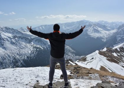 A man stands on a snowy mountain peak with arms raised, embracing the breathtaking view.